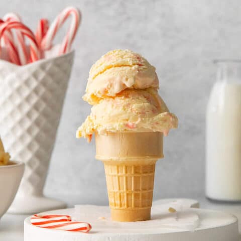 Two scoops of peppermint ice cream in an ice cream cone with a white container of candy canes in the background and more scoops of ice cream on either side.