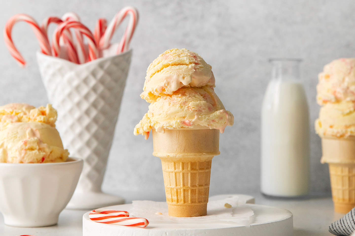 Two scoops of peppermint ice cream in an ice cream cone with a white container of candy canes in the background and more scoops of ice cream on either side.