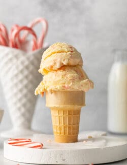 Two scoops of peppermint ice cream in an ice cream on a white marble round, with a ceramic cone of candy canes in the background.