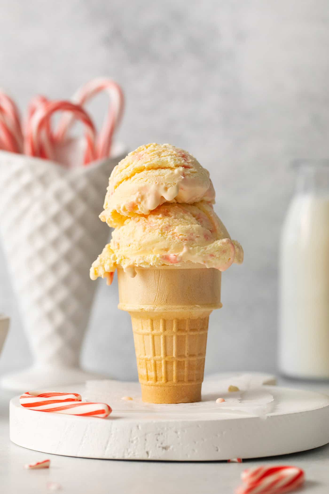 Two scoops of peppermint ice cream in an ice cream on a white marble round, with a ceramic cone of candy canes in the background.