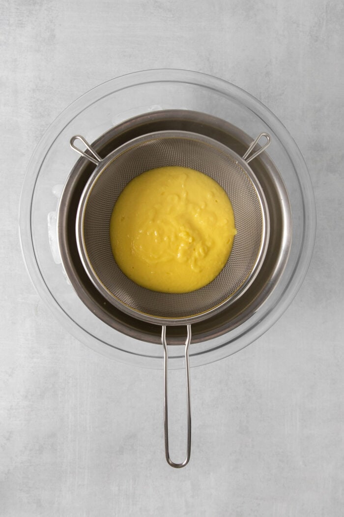 A yellowish ice cream base is poured through a metal strainer set over a metal bowl.