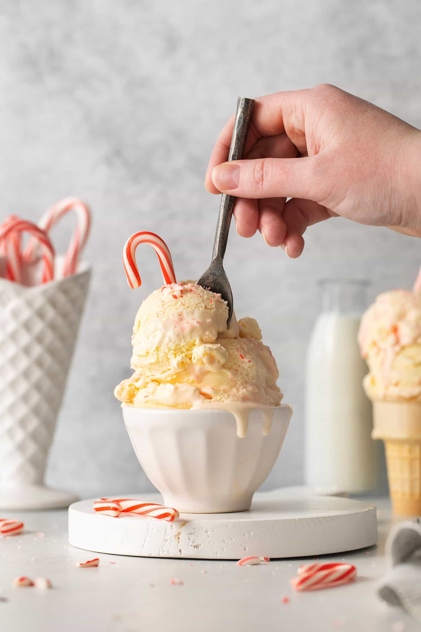 A hand holding a spoon digs into a bowl containing two scoops of peppermint ice cream topped by a candy cane.