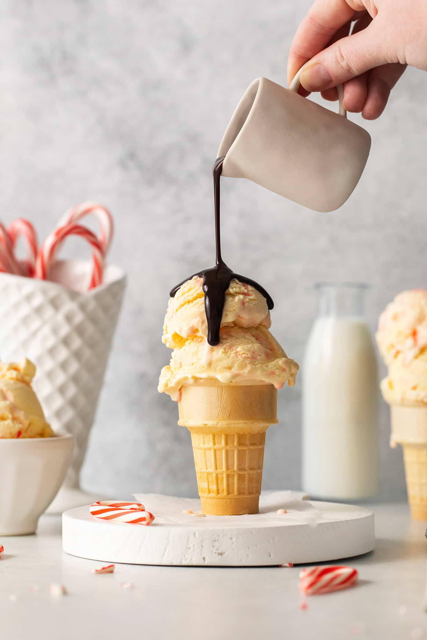 A hand holding a white pitcher pours chocolate sauce over an ice cream cone full of two scoops of peppermint ice cream, with candy canes in the background.