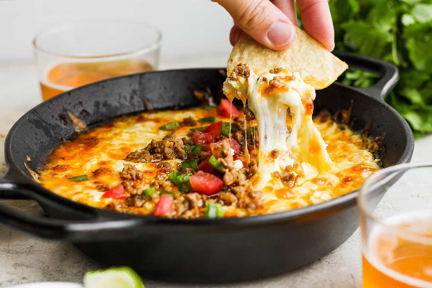 A hand holding a corn chip dips into a bowl of tomato-topped queso fundido.