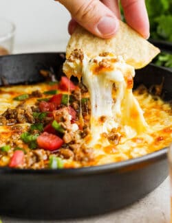 A hand holding a corn chip dips into a bowl of tomato-topped queso fundido.