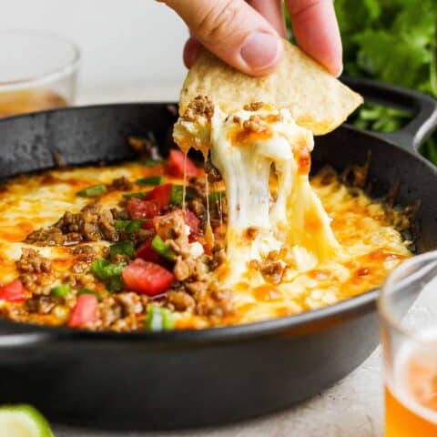 A hand holding a corn chip dips into a bowl of tomato-topped queso fundido.