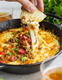 A hand holding a corn chip dips into a bowl of tomato-topped queso fundido with melted cheese and chorizo.