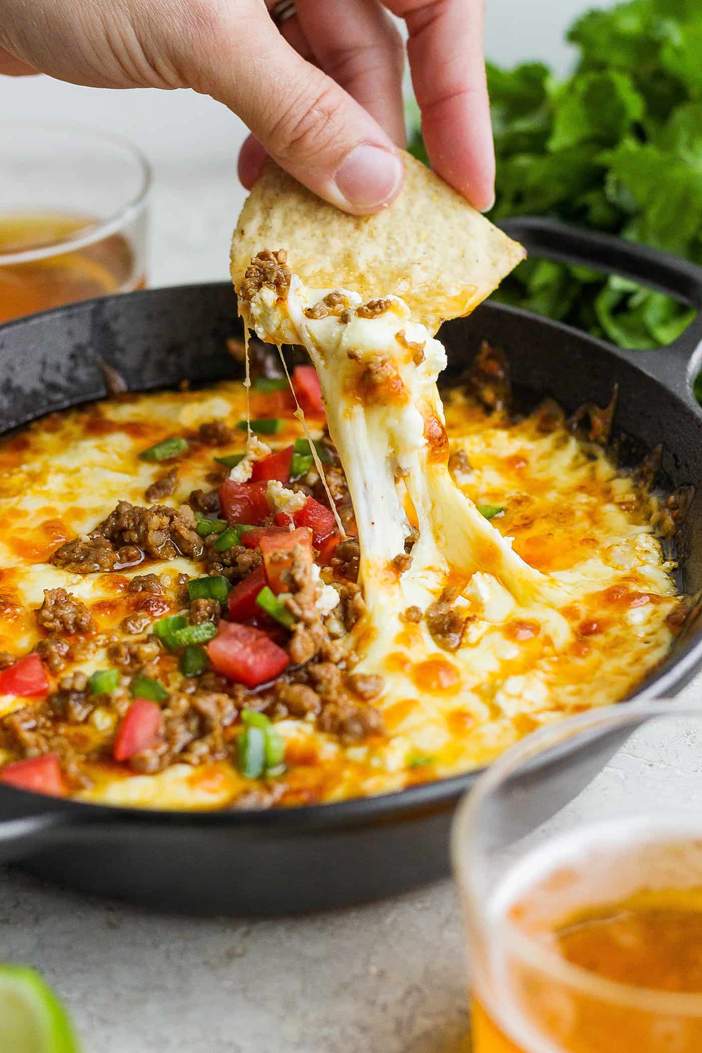 A hand holding a corn chip dips into a bowl of tomato-topped queso fundido with melted cheese and chorizo.