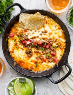 A black cast iron skillet of queso fundido topped with chorizo, tomatoes, and jalapeno and garnished with tortilla chips, with lime wedges on the side.