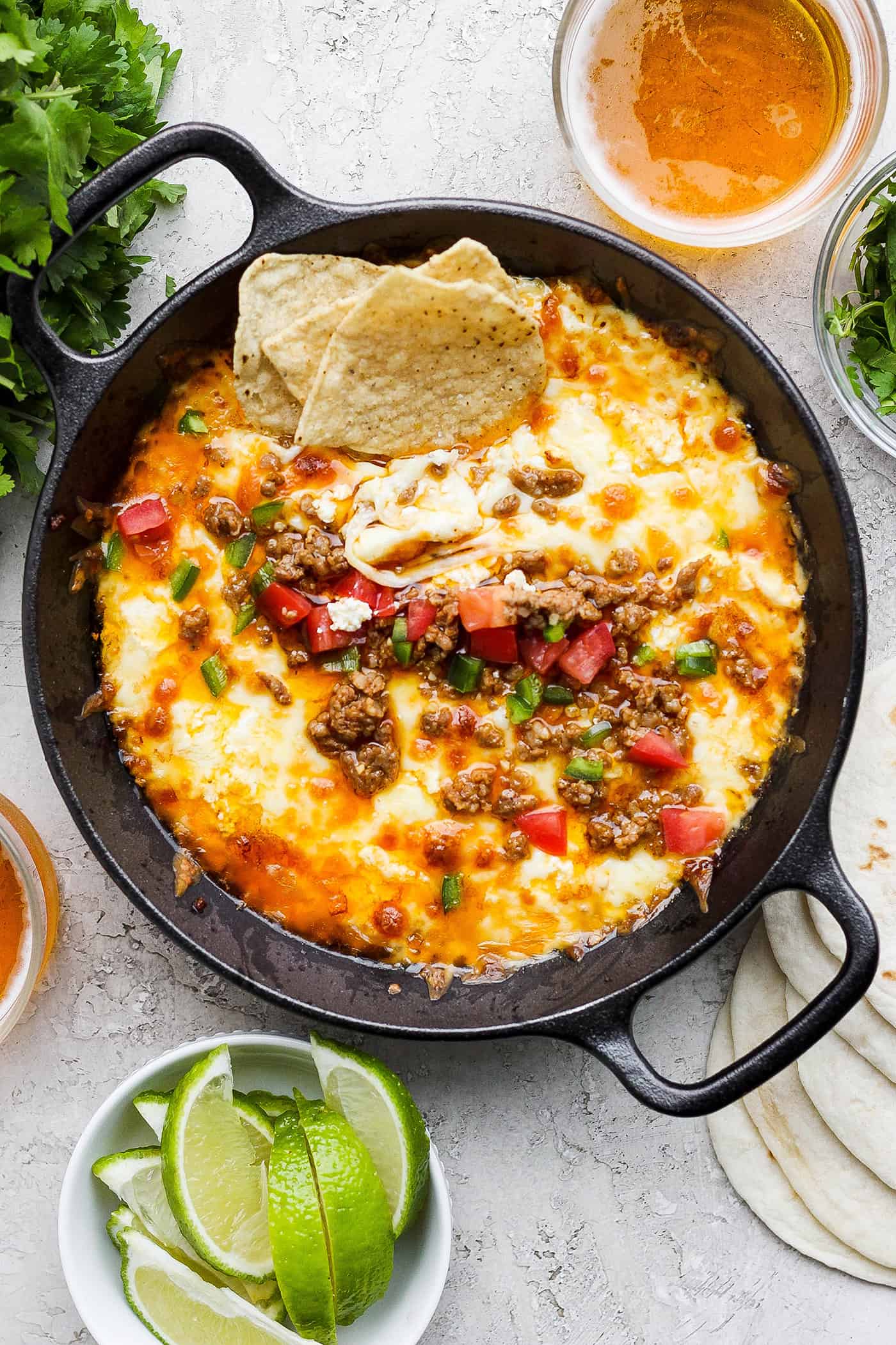 A black cast iron skillet of queso fundido topped with chorizo, tomatoes, and jalapeno and garnished with tortilla chips, with lime wedges on the side.