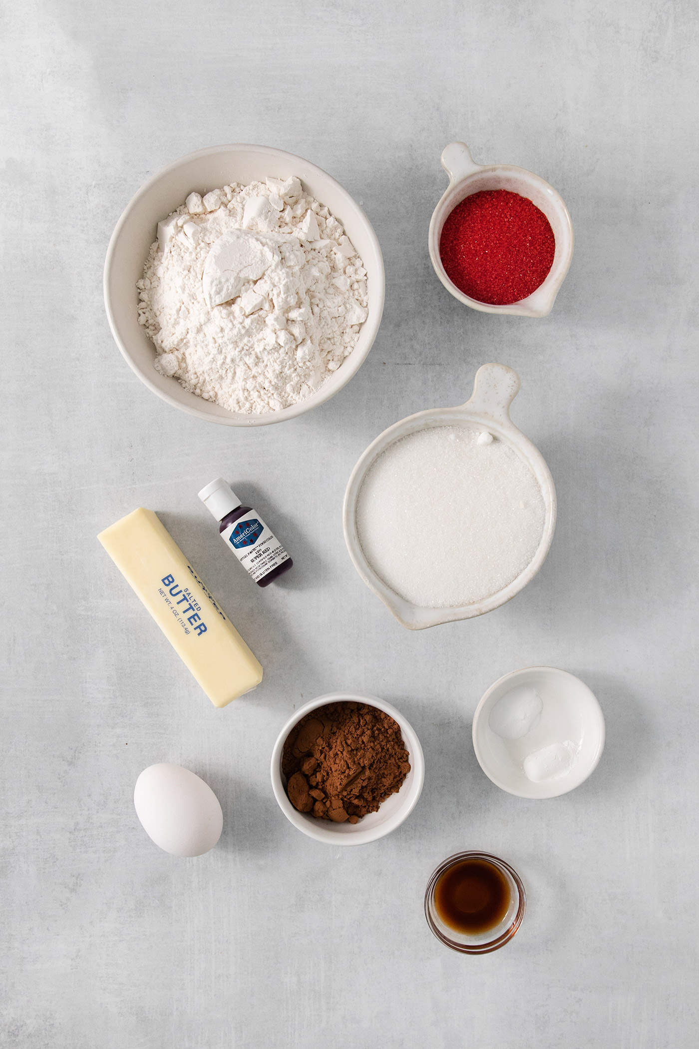 The ingredients to make red velvet cookies are shown portioned out: butter, flour, milk, red food coloring, egg, cocoa powder, vanilla.