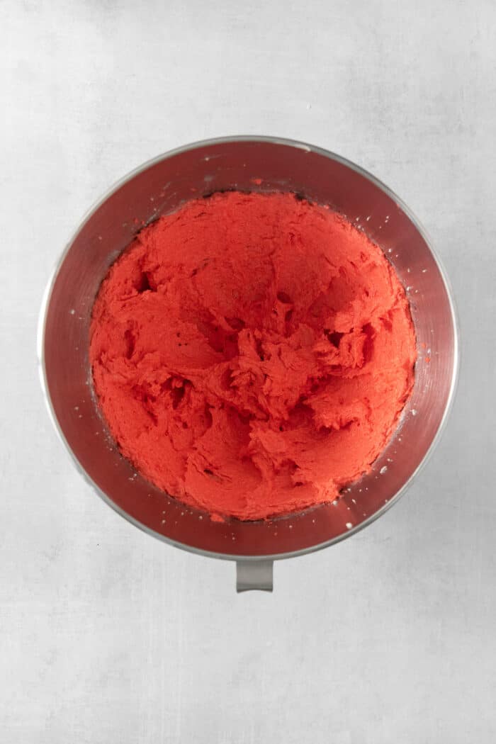 Red food coloring is adding to butter and sugar in a metal bowl.