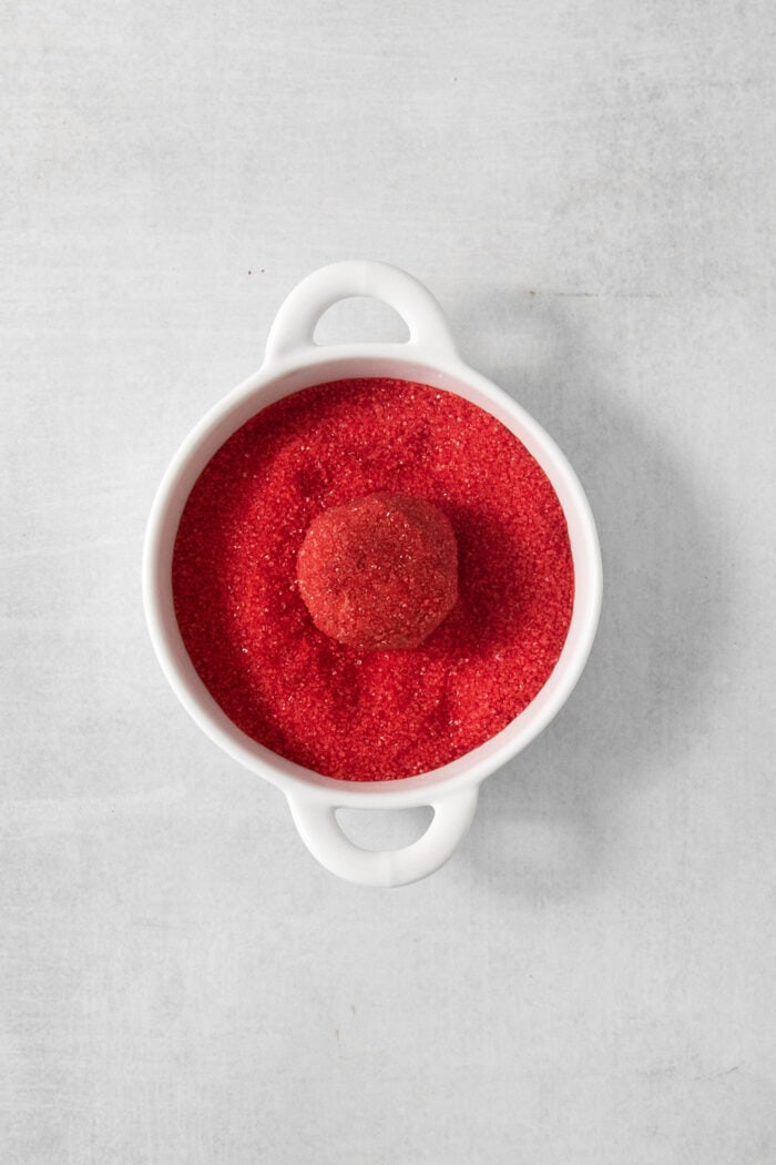 A red velvet cookie is placed in a bowl of red sanding sugar.