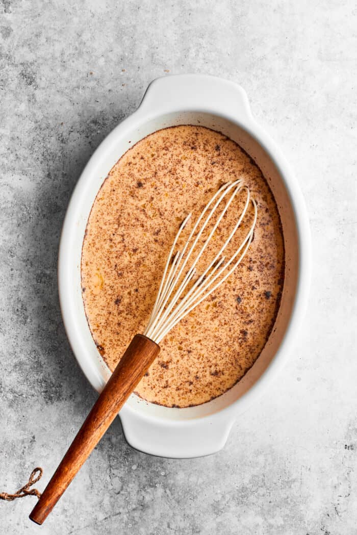 A whisk blends together the egg batter for bananas foster French toast in a white baking dish.