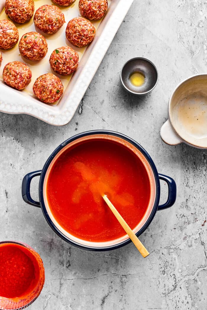A wooden spoon stirs a pot of tomato sauce with uncooked porcupine meatballs to its left.