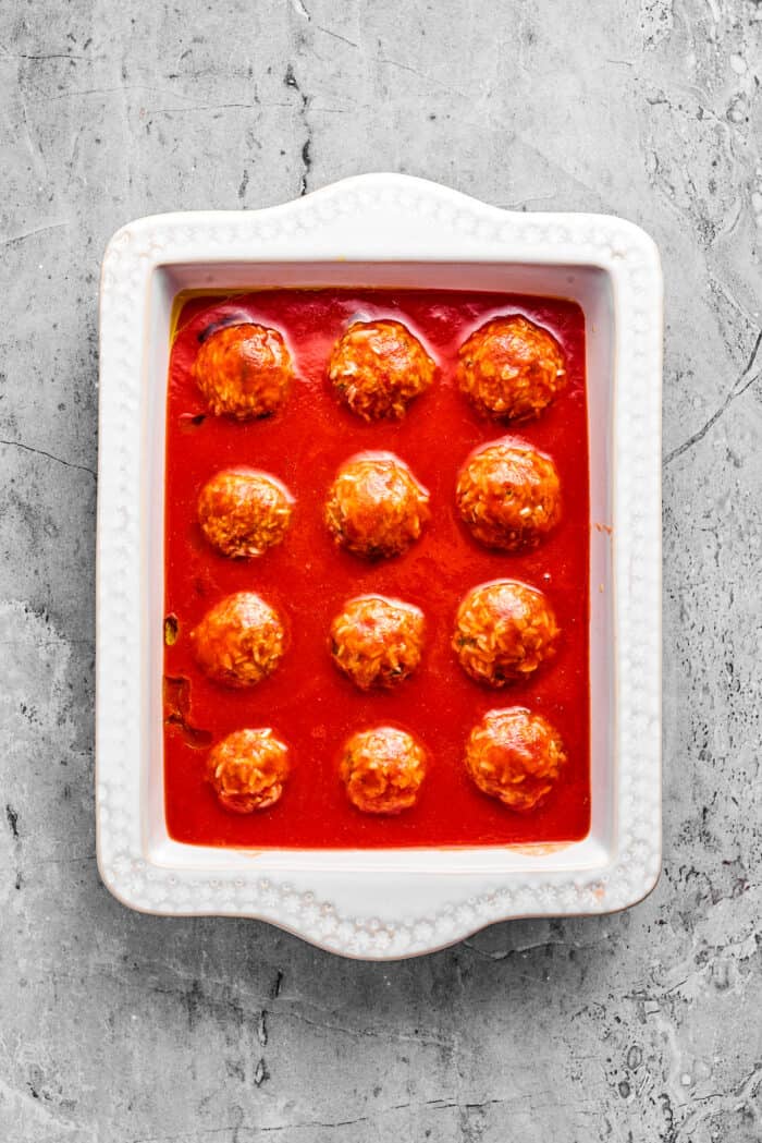 Meatballs are placed in red sauce in a white baking dish.