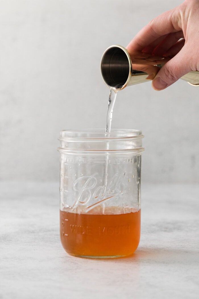 A hand pours simple syrup juice into a mason jar to make an amaretto sour.