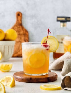 A lemon-garnished glass of amaretto sour is shown on a wooden circle with lemon slices around it and lemons in the background.