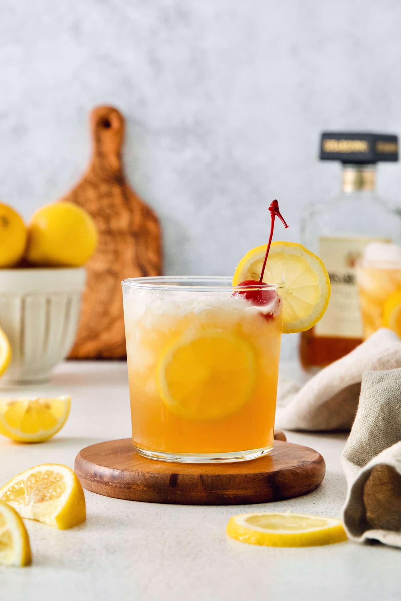 A lemon-garnished glass of amaretto sour is shown on a wooden circle with lemon slices around it and lemons in the background.