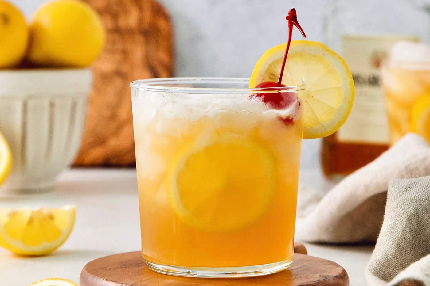 A glass of amaretto sour is garnished with a cherry and a slice of lemon on a wooden circle.