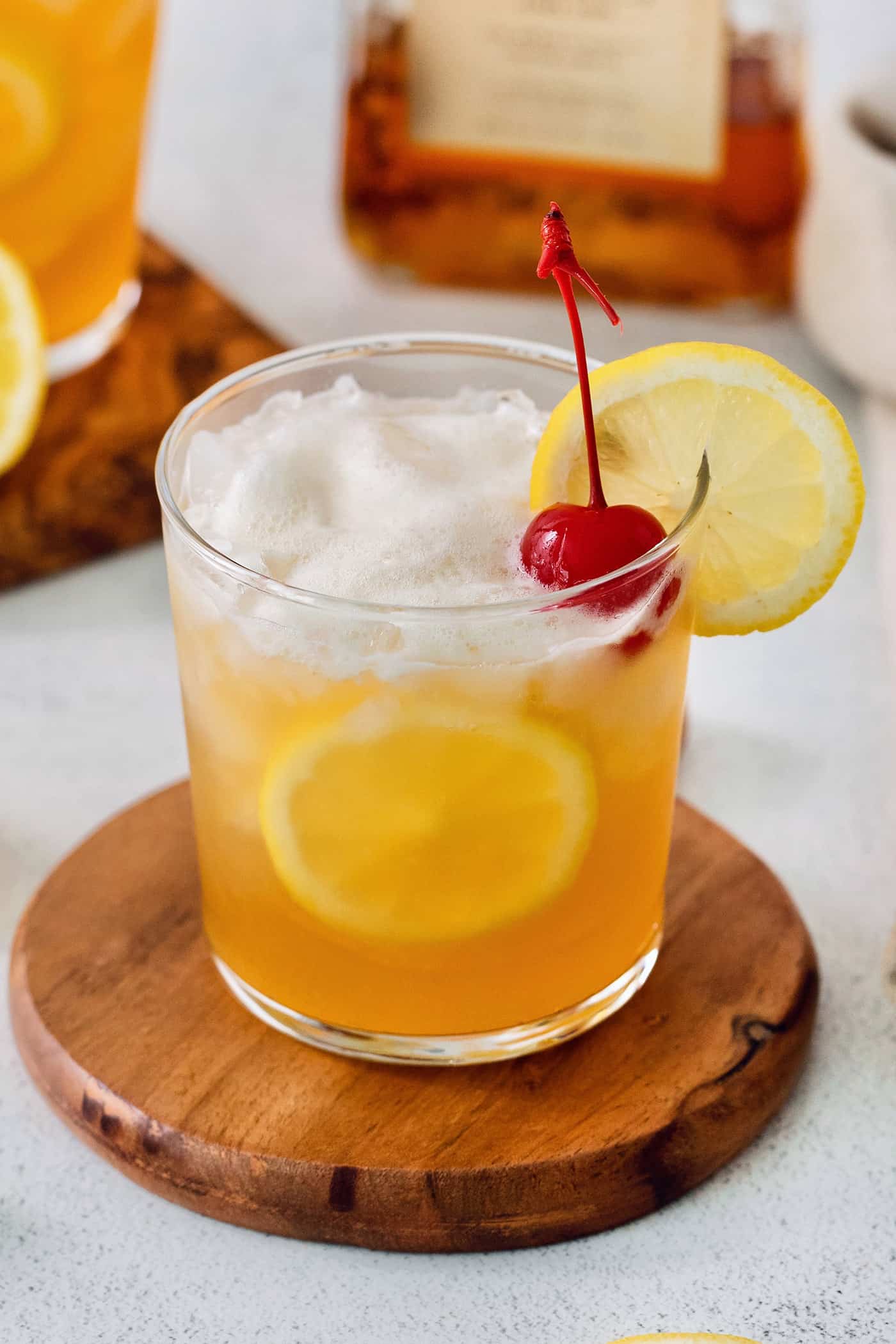 A glass of amaretto sour is garnished with a cherry and a slice of lemon on a wooden circle.