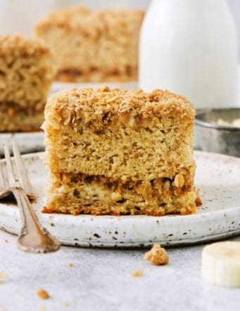 A piece of banana coffee cake on a white plate with a fork, with more pieces of cake shown in the background.