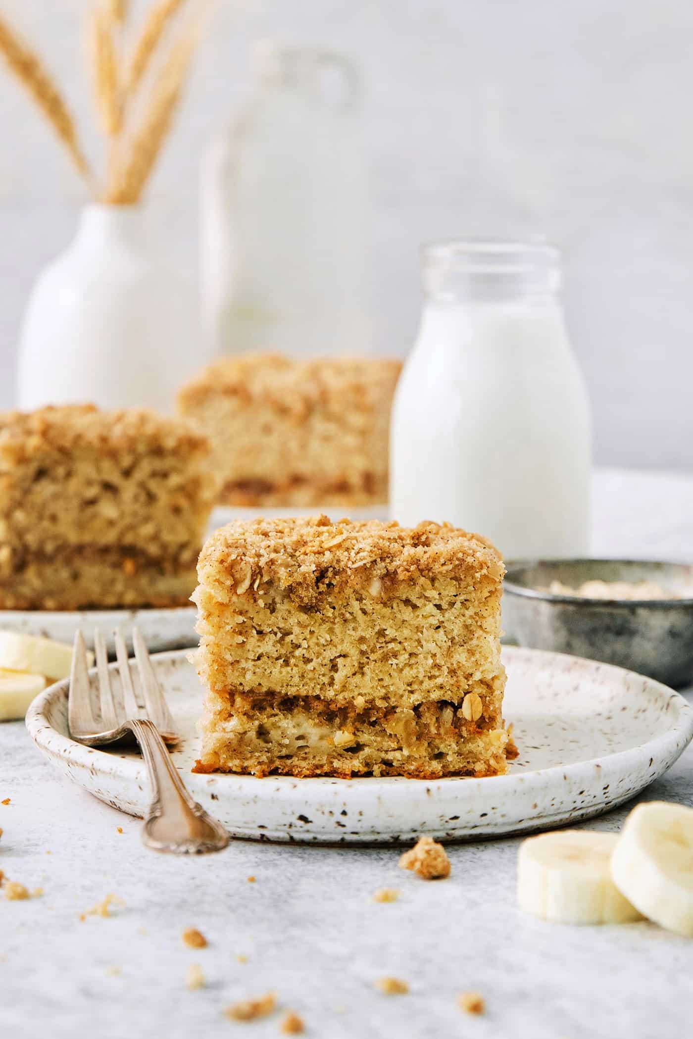 A piece of banana coffee cake on a white plate with a fork, with more pieces of cake shown in the background.