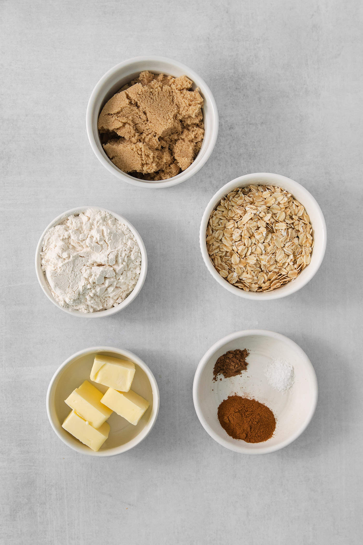 The ingredients for banana coffee cake streusel are shown portioned out in bowls: butter, flour, oats, brown sugar, salt, cinnamon, and nutmeg.