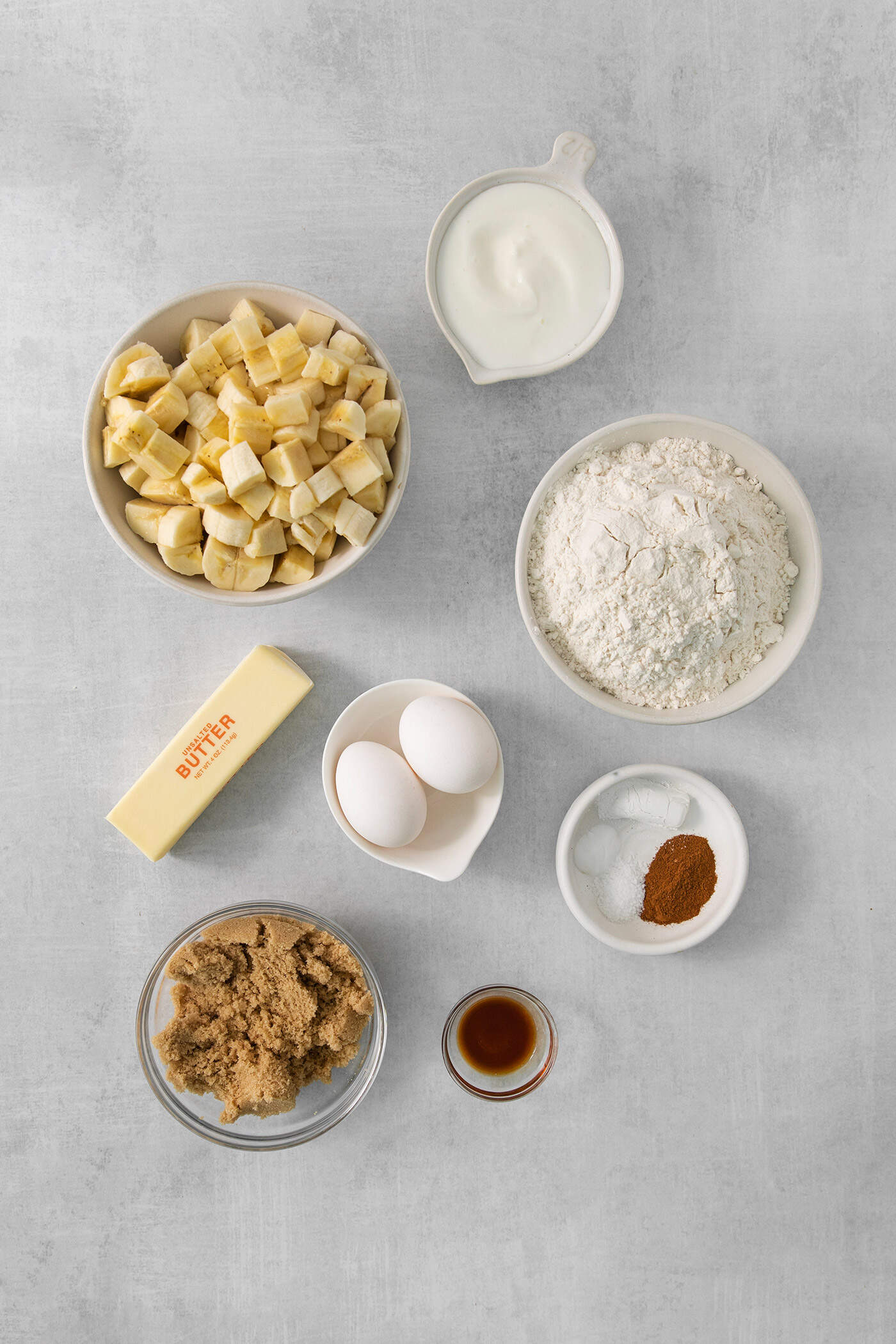 The ingredients for banana coffee cake are shown portioned out in bowls: butter, flour, eggs, vanilla, brown sugar, sour cream, cinnamon, baking powder, baking soda, and salt, and bananas,