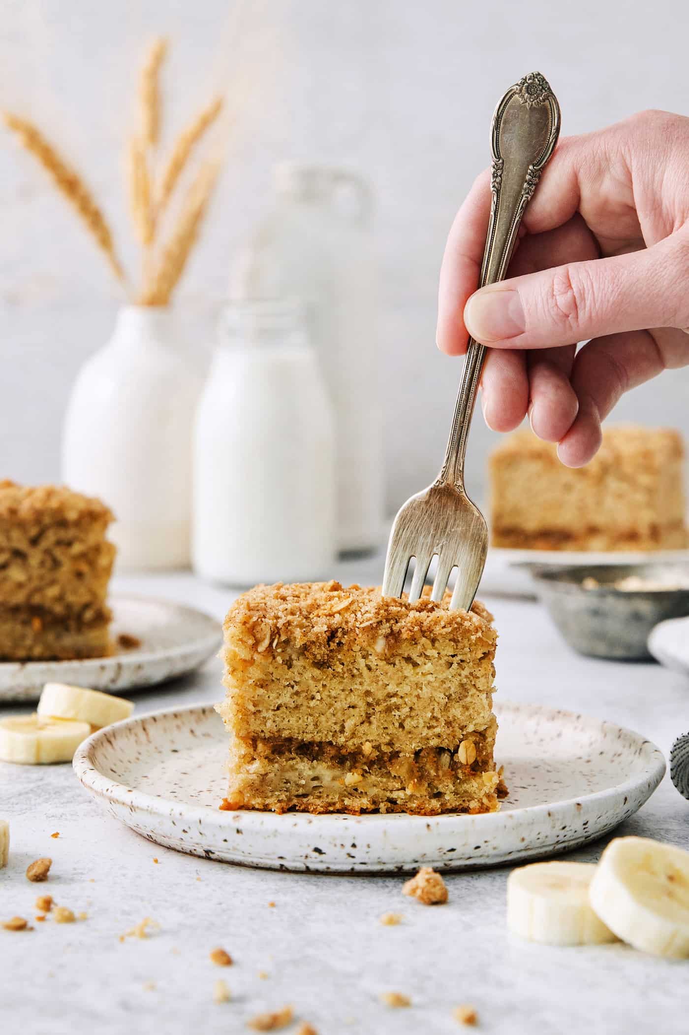 A hand holding a fork cuts into a thick slice of banana coffee cake on a white plate.