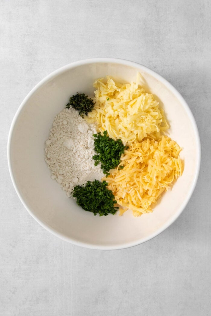 Cheddar cheese, gouda, and pimento cheese plus bisquick and fresh herbs are placed in a white bowl.