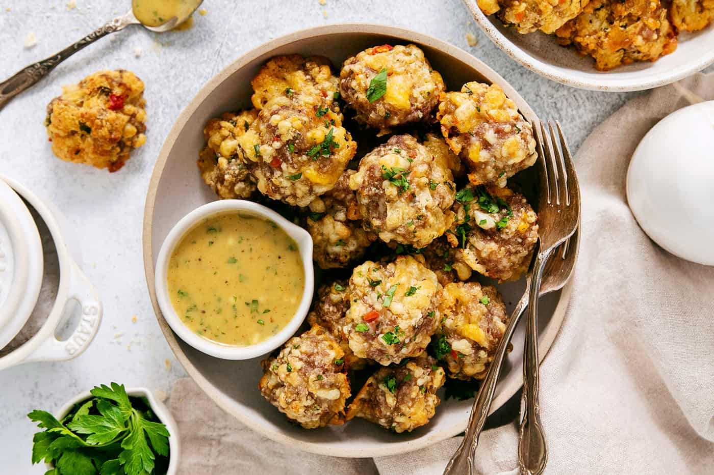 A big white bowl with serving utensils holds sausage cheese balls with a bowl of yellow dipping sauce nestled against the balls.