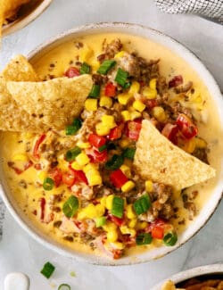 A white bowl of cheesy corn sausage dip is garnished with Rotel and corn chips.