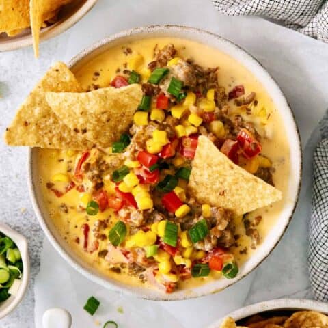 A white bowl of cheesy corn sausage dip is garnished with Rotel and corn chips.