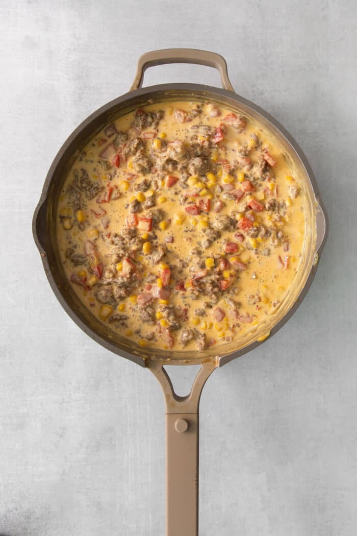 Corn and Rotel tomatoes are added to a skillet full of cooked sausage and melted cheese.