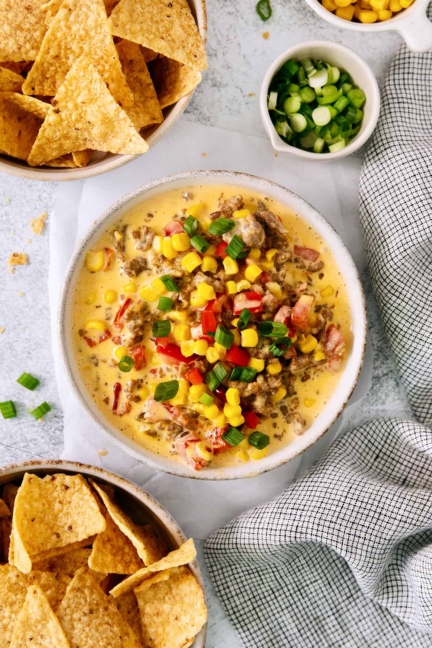 Cheesy corn sausage dip is topped with tomatoes, green onions, and tomatoes with bowls of corn chips for dipping nearby.