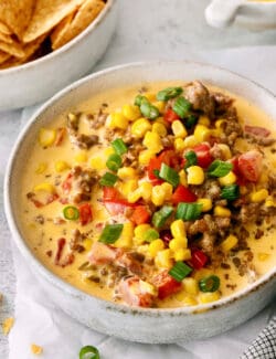 A bowl of cheesy corn sausage dip is topped with corn, tomatoes, and green onions.