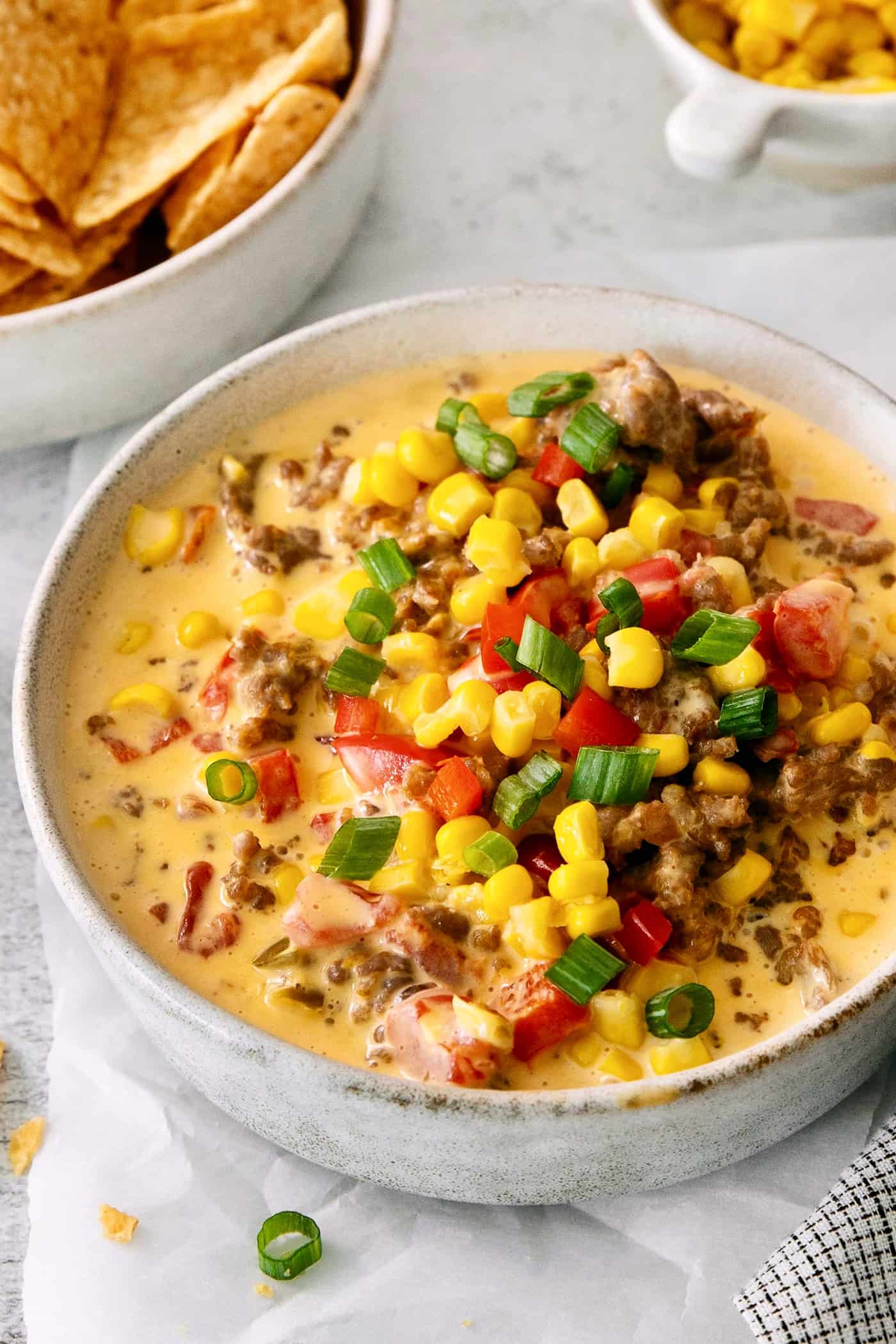 A bowl of cheesy corn sausage dip is topped with corn, tomatoes, and green onions.