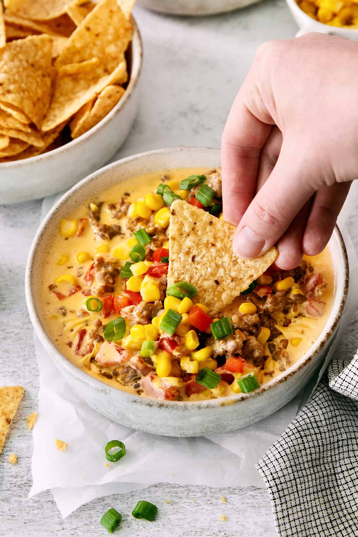 A hand dips a corn chip into a bowl of cheesy corn sausage dip.