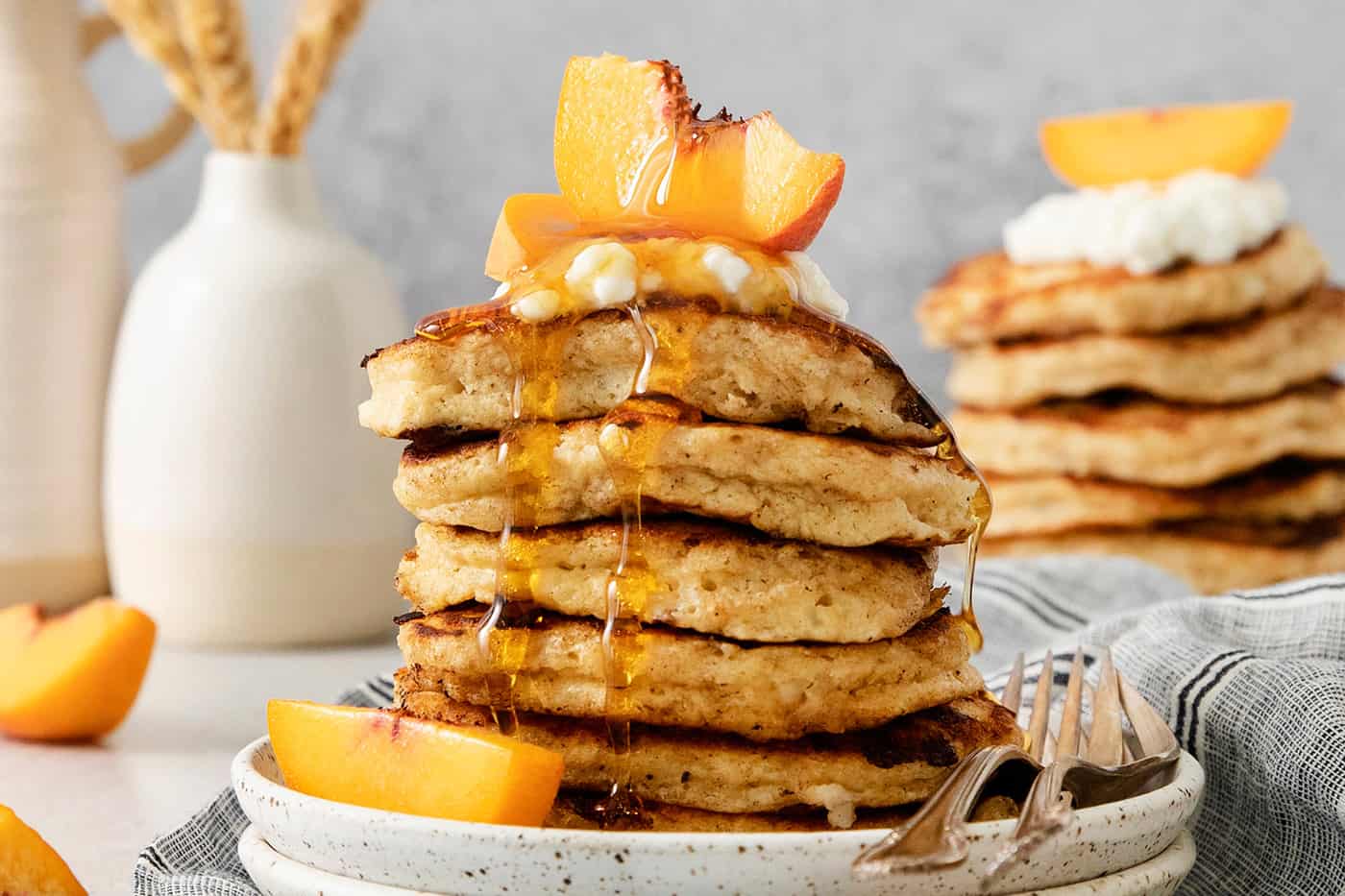 A tall stack of cottage cheese pancakes on a plate is topped with sliced fresh peaches and maple syrup.