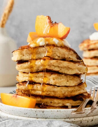A tall stack of cottage cheese pancakes on a plate is topped with sliced fresh peaches and maple syrup.