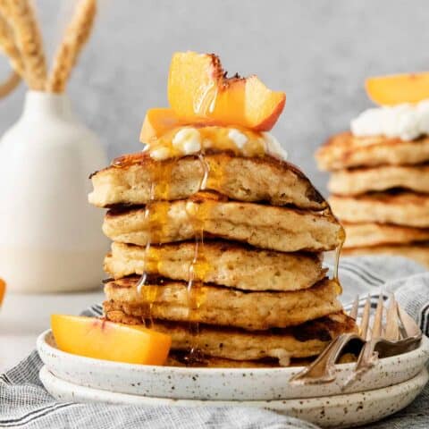 A tall stack of cottage cheese pancakes on a plate is topped with sliced fresh peaches and maple syrup.