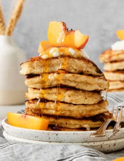 A tall stack of cottage cheese pancakes on a plate is topped with sliced fresh peaches and maple syrup.