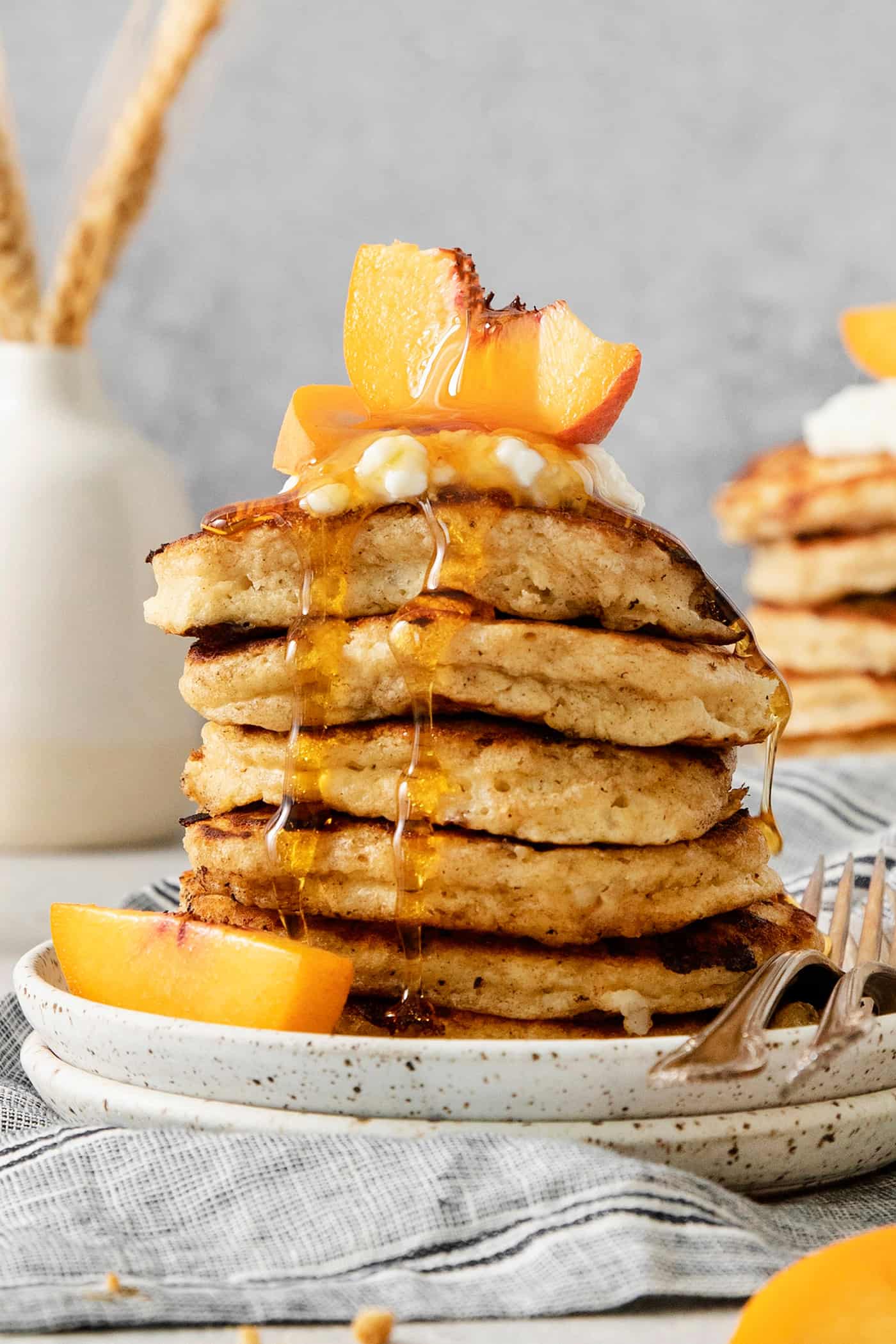 A tall stack of cottage cheese pancakes on a plate is topped with sliced fresh peaches and maple syrup.