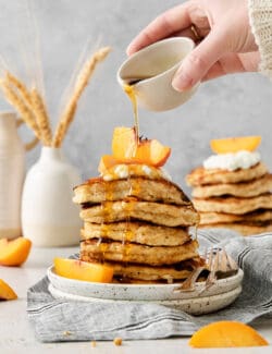 A hand drizzles maple syrup from a small white pitcher over a stack of cottage cheese pancakes.