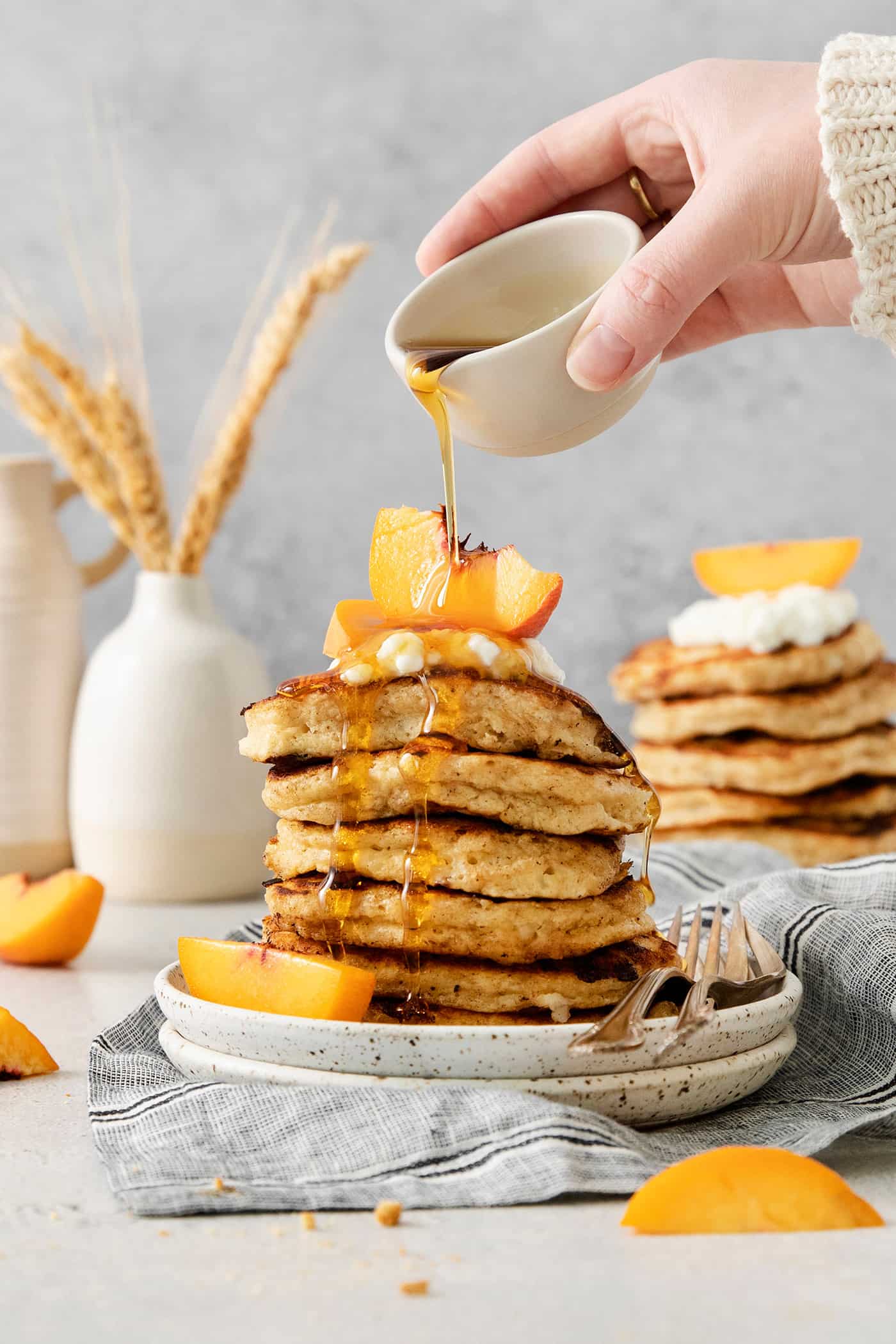 A hand drizzles maple syrup from a small white pitcher over a stack of cottage cheese pancakes.