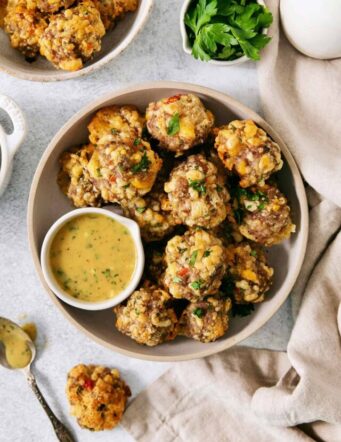 A big white bowl with serving utensils holds sausage cheese balls with a bowl of yellow dipping sauce nestled against the balls.