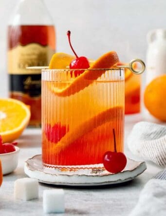 A bottle of brandy is seen in the background with a glass of brandy old fashioned in the foreground.