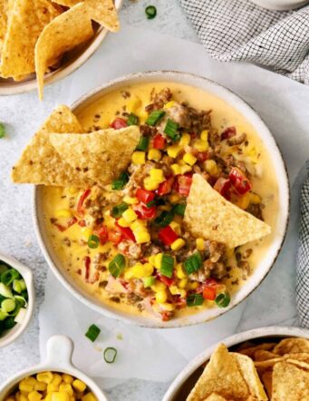 A white bowl of cheesy corn sausage dip is garnished with Rotel and corn chips.