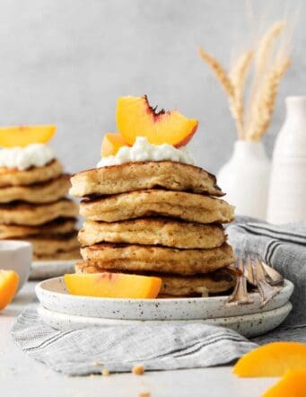 Peach slices and whipped cream top cottage cheese pancakes with more seen in the background.